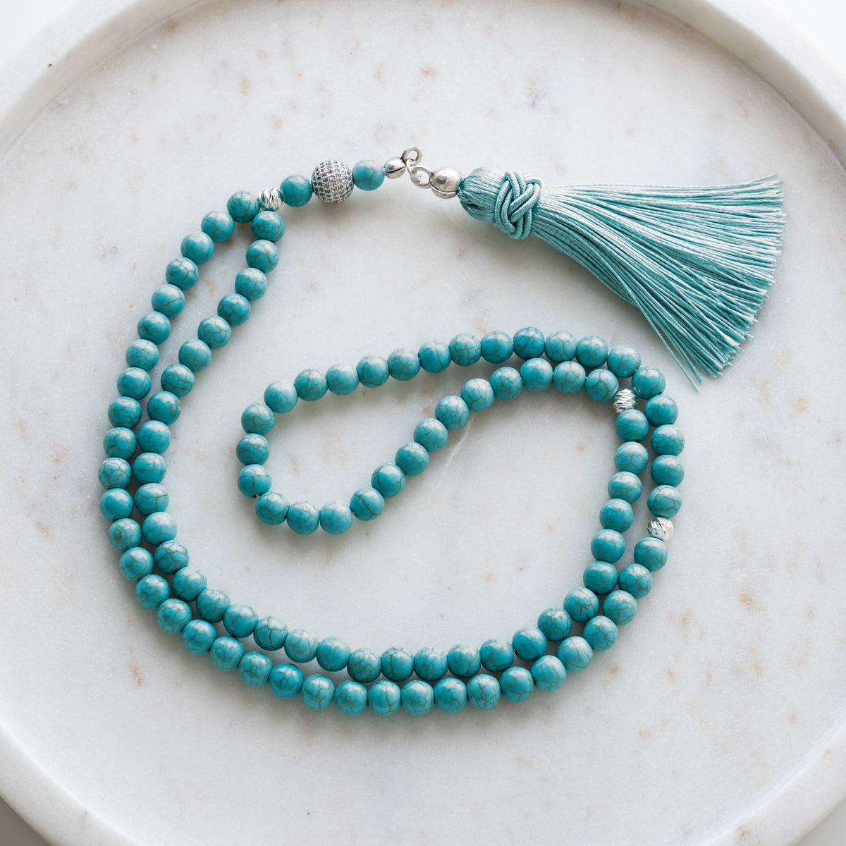 Tasbih (Misbaha) aus Türkis mit 99 Natursteinperlen, silbernen Zwischenperlen und türkisfarbener Quaste auf weißem Marmor